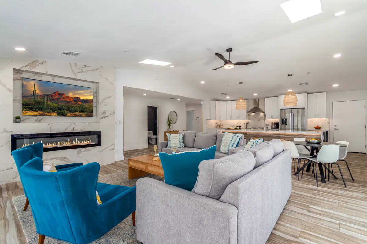 A spacious open-concept living area with a sofa seating arrangement facing a wall-mounted TV (displaying landscape art), a modern electric fireplace, and the kitchen area visible in the background. The room is filled with natural light, and the furnishings look plush and inviting, perfect for gatherings and relaxation.