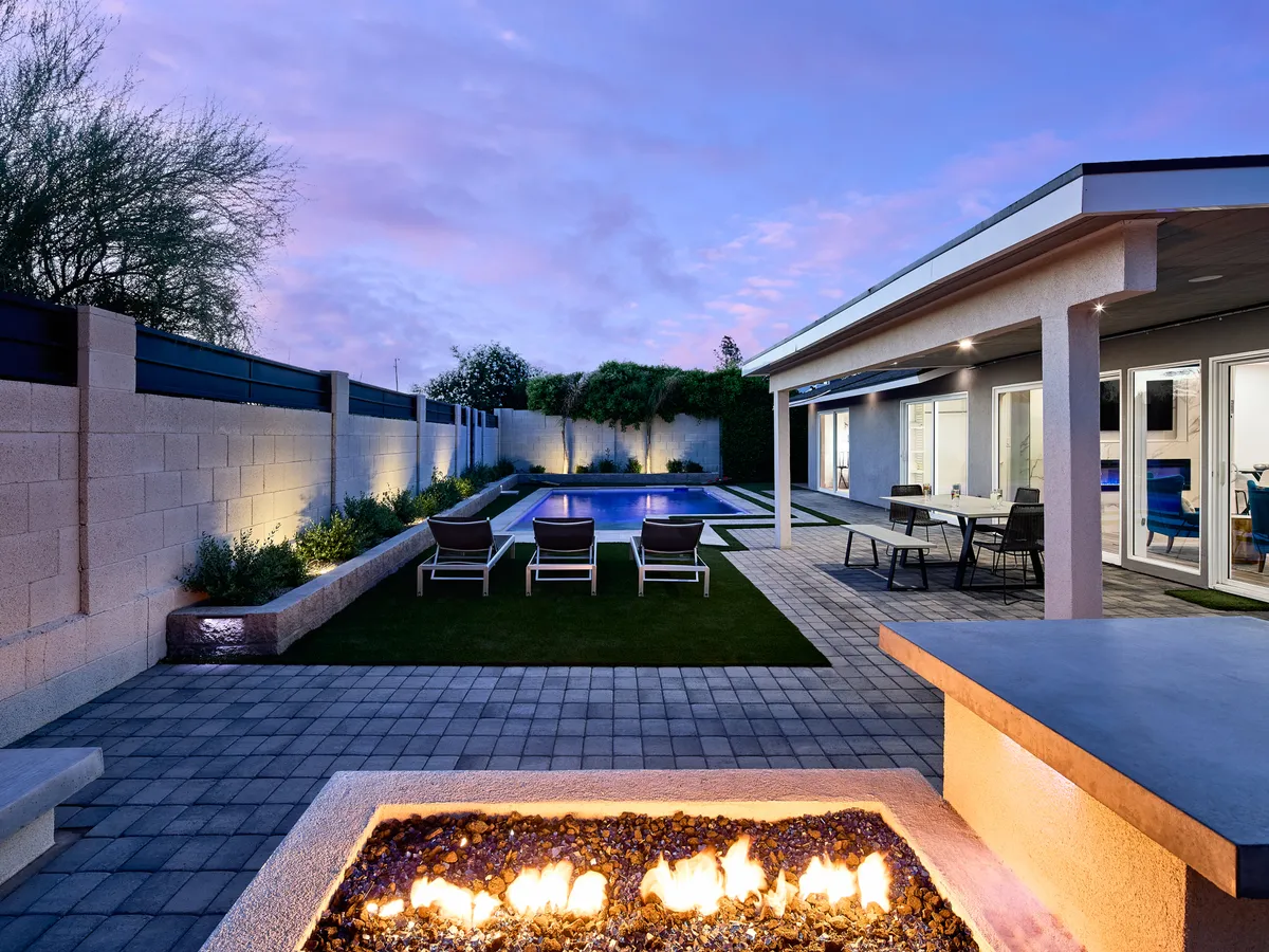 A serene backyard scene featuring a sparkling swimming pool, a modern fire pit surrounded by cozy patio seating, and lush greenery providing privacy and ambiance. The space is designed for both relaxation and social gatherings, with multiple areas to enjoy the outdoors day or night.