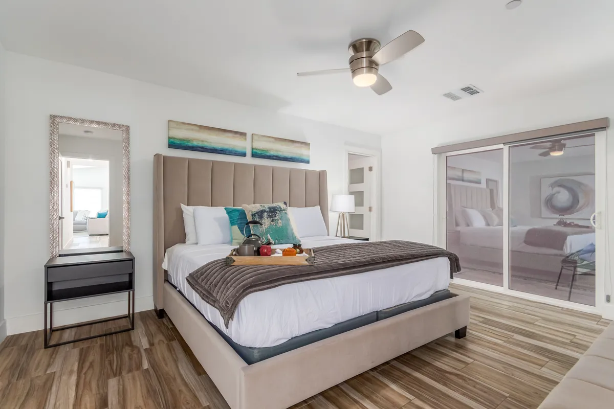A serene bedroom featuring a king-sized bed with a tufted headboard, soft linens, a decorative mirror, a fruit tray on the bed, and calming artwork above the bed. Sunlight filters in, creating a peaceful ambiance ideal for relaxation.