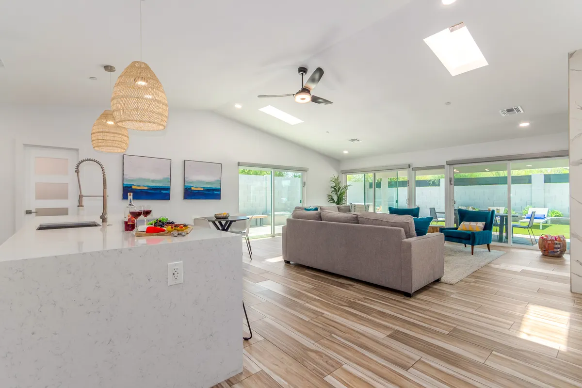 A modern interior view showcasing a kitchen island with bar stools, a comfortable sofa, large windows letting in abundant sunlight, and contemporary artwork adorning the wall. The space flows naturally between the kitchen and living area, emphasizing the open-concept design.