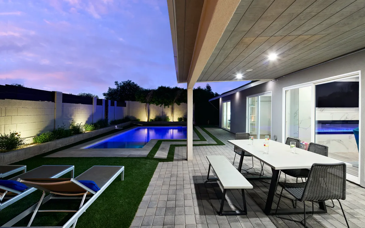 A tranquil backyard scene with a sparkling swimming pool, outdoor dining table, and lounge chairs surrounded by lush greenery. The setting is perfect for cooling off, relaxing, or socializing after a day outdoors.