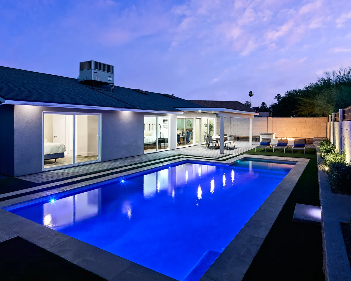 A serene backyard scene at sunset featuring a sparkling swimming pool surrounded by a landscaped yard. There are comfortable lounge chairs and an inviting outdoor seating area adjacent to large glass sliding doors, creating a seamless transition between indoor and outdoor living. The atmosphere suggests relaxing swims, starlit dinners, and gathering spaces ideal for groups.
